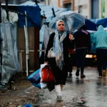 أغرقت الأمطار الغزيرة الناجمة عن منخفض جوي جديد مستشفى الشفاء وآلاف الخيام التي تؤوي نازحين، وسط تحذيرات من تفشي الأوبئة. وقد أعلنت وزارة الصحة في غزة وفاة طفل رضيع بسبب انخفاض حاد في درجات حرارة الجسم جراء البرد القارس. من جهة ثانية أفاد مراسل الأناضول بأن مياه الأمطار تسربت إلى أقسام في مجمع الشفاء الطبي بمدينة غزة، وخاصة قسم الاستقبال والطوارئ، مما أدى إلى تعطيل العمل فيه. ومجمع الشفاء الطبي أكبر مستشفيات القطاع وقد تعرض لدمار هائل وعمليات قصف وحرق لجميع مبانيه خلال الإبادة الإسرائيلية في غزة. وعملت وزارة الصحة بغزة على ترميم بعض المباني بالمستشفى خلال الشهرين الماضيين بعد دخول اتفاق لوقف إطلاق النار حيز التنفيذ في 10 أكتوبر/تشرين الأول 2025، لكن حجم الأضرار الكبيرة ونقص الإمكانات يحولان دون عودة العمل فيه بصورة طبيعية، خاصة مع عرقلة إسرائيل دخول المستلزمات والأجهزة الطبية والأدوية. A displaced Palestinian woman holds an umbrella to shield from the rain in Gaza City on December 15, 2025. قطاع غزة يشهد منخفضا جويا جديدا يفاقم معاناة النازحين (الفرنسية) أمطار ورياح عاتية وفي سياق متصل، قال شهود عيان لمراسل الأناضول، إن آلافا من خيام النازحين أغرقتها مياه الأمطار وتطايرت بفعل الرياح الشديدة التي تضرب القطاع منذ مساء الاثنين. وذكر الشهود أن آلاف الفلسطينيين استيقظوا فجر الثلاثاء، وقد أغرقت مياه الأمطار خيامهم أو تسببت الرياح بتطايرها مع مقتنياتهم. وقال الفلسطيني خالد عبد العزيز: استيقظنا على صوت رياح عاتية تضرب خيمتنا وحاولنا تثبيتها وإمساكها بأيدينا لكن الرياح اقتلعتها وتطايرت كل مقتنياتنا. وأضاف عبد العزيز: أجلس مع زوجتي وأطفالي في العراء حاليا تحت مياه الأمطار. لا يوجد أي مكان نلجأ إليه. في حين كانت الفلسطينية مها أبو جزر تركض مع أطفالها الثلاثة دون تحديد وجهة، بعد أن أغرقت مياه الأمطار خيمتها بصورة كاملة في منطقة المواصي غربي مدينة خان يونس، جنوبي القطاع. إعلان وحاول مئات الفلسطينيين الاحتماء من مياه الأمطار تحت أجزاء من مبان دمرها الجيش الإسرائيلي في مدينة غزة، حسب الشهود. A Palestinian family rides on a cart amid the rain in Gaza City, Monday, Dec. 15, 2025. (AP Photo/Jehad Alshrafi) آلاف الفلسطينيين استيقظوا فجر الثلاثاء، وقد أغرقت مياه الأمطار خيامهم أو تسببت الرياح بتطايرها مع مقتنياتهم (أسوشيتد برس) في السياق، أعلن الدفاع المدني في غزة عن إصابات في انهيار منزل في شارع الشفاء غربي مدينة غزة. وطالب المتحدث باسم بلدية غزة للجزيرة بالضغط على الاحتلال الإسرائيلي من أجل فتح المعابر وإدخال المساعدات. من جهته حذر متحدث الدفاع المدني في غزة محمود بصل، من أن آلاف المنازل التي دمرت جزئيا خلال الإبادة الإسرائيلية مهددة بالانهيار في أي لحظة بفعل الأمطار والرياح العاتية. وقال بصل لمراسل الأناضول: "هذه المنازل تشكل خطرا جسيما على حياة مئات آلاف الفلسطينيين الذين لم يجدوا أي ملجأ وقد حذرنا العالم مرارا ولكن دون جدوى". وخلال المنخفض الأول، أفادت معطيات رسمية بانهيار 13 مبنى على الأقل، من المباني المتضررة جراء الإبادة الإسرائيلية، وذلك على رؤوس ساكنيها الذين لجؤوا إليها للاحتماء من الأمطار والبرد. يأتي ذلك وسط تنصل إسرائيل من الإيفاء بالتزاماتها التي نص عليها اتفاق وقف إطلاق النار الساري منذ 10 أكتوبر/تشرين الأول الماضي، وبروتوكوله الإنساني، بما فيه إدخال مواد الإيواء و300 ألف خيمة وبيت متنقل، وفق ما أكده مرارا المكتب الإعلامي الحكومي بغزة. تحذيرات من أمراض كما حذر المتحدث باسم منظمة اليونيسيف في فلسطين جوناثان كريكس من انتشار الأمراض بين الأطفال في قطاع غزة نتيجة الظروف الجوية الباردة والماطرة التي يمر بها القطاع. وقال كريكس للجزيرة إن حجم الاحتياجات كبير جدا. وفي سياق متصل، جددت وكالة الأمم المتحدة لإغاثة وتشغيل اللاجئين الفلسطينيين ( الأونروا) تحذيرها من تدهور أوضاع الغزيين، بسبب قلة المساعدات الإغاثية ومستلزمات الإيواء التي تسمح إسرائيل بإدخالها إلى القطاع. وقال عدنان أبو حسنة، المستشار الإعلامي للأونروا، في مقابلة مع الجزيرة إن حوالي مليوني فلسطيني يتكدسون في نحو 80 كيلومترا مربعا، في حين تحتل إسرائيل نصف القطاع تقريباً، وينتشر الركام في المساحة المتبقية، في ظل انتشار متزايد للأمراض والأوبئة. وفي هذه الأثناء بدأ الدفاع المدني في القطاع انتشال جثامين عشرات الشهداء، بينهم أطفال ونساء، من بين أنقاض مبنى يؤوي نازحين في حي الرمال غربي مدينة غزة، وتتم هذه العمليات المعقدة بما توفَّر من معدات وآليات نجت من شهور الحرب الطويلة. وإلى جانب تلك الأزمات التي يعيشها القطاع، تواصل قوات الاحتلال الإسرائيلي تنفيذ غارات وقصف مدفعي وإطلاق نار مكثفا من الدبابات في مناطق انتشارها بحي التفاح شرقي مدينة غزة. كما أفاد مراسل الجزيرة بأن مناطق شرقي مدينتي رفح وخان يونس وكذلك شرقي البريج تعرضت لغارات جوية وقصف مدفعي إسرائيلي في مناطق انتشار جيش الاحتلال. وخلّفت حرب الإبادة الإسرائيلية في غزة التي بدأت في 8 أكتوبر/تشرين الأول 2023 بدعم أميركي واستمرت سنتين، أكثر من 70 ألف شهيد وما يفوق 171 ألف جريح، معظمهم أطفال ونساء، ودمارا طال 90% من البنى التحتية المدنية.